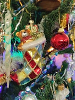 A colorful Christmas tree decorated with an ornate stocking ornament featuring a teddy bear. The stocking has a patchwork pattern of red, blue, and gold squares with glittery accents. Next to it is a shiny red bauble and other decorations such as a transparent icicle and gold tinsel. The tree is lush with green needles and surrounded by glowing Christmas lights.