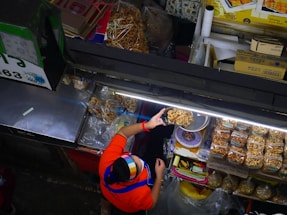 A vendor wearing a colorful headscarf and a red shirt serves dried snacks at a market stall. The display includes bags of packaged snacks and individually served items, neatly arranged under bright fluorescent lights. Various items and boxes are organized around the stall, showcasing a busy and organized market setup.