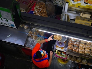 A vendor wearing a colorful headscarf and a red shirt serves dried snacks at a market stall. The display includes bags of packaged snacks and individually served items, neatly arranged under bright fluorescent lights. Various items and boxes are organized around the stall, showcasing a busy and organized market setup.