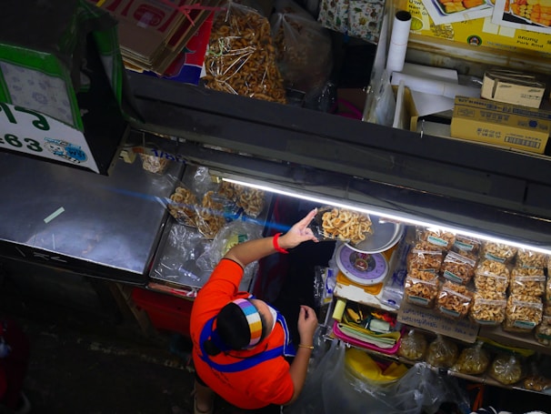 A vendor wearing a colorful headscarf and a red shirt serves dried snacks at a market stall. The display includes bags of packaged snacks and individually served items, neatly arranged under bright fluorescent lights. Various items and boxes are organized around the stall, showcasing a busy and organized market setup.