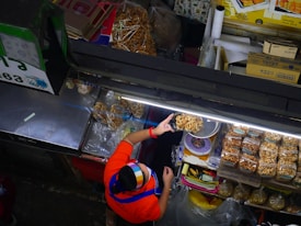 A vendor wearing a colorful headscarf and a red shirt serves dried snacks at a market stall. The display includes bags of packaged snacks and individually served items, neatly arranged under bright fluorescent lights. Various items and boxes are organized around the stall, showcasing a busy and organized market setup.