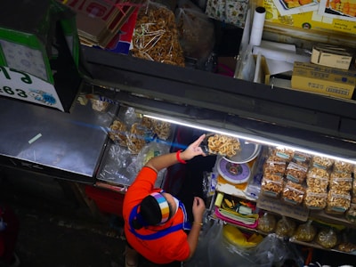 A vendor wearing a colorful headscarf and a red shirt serves dried snacks at a market stall. The display includes bags of packaged snacks and individually served items, neatly arranged under bright fluorescent lights. Various items and boxes are organized around the stall, showcasing a busy and organized market setup.