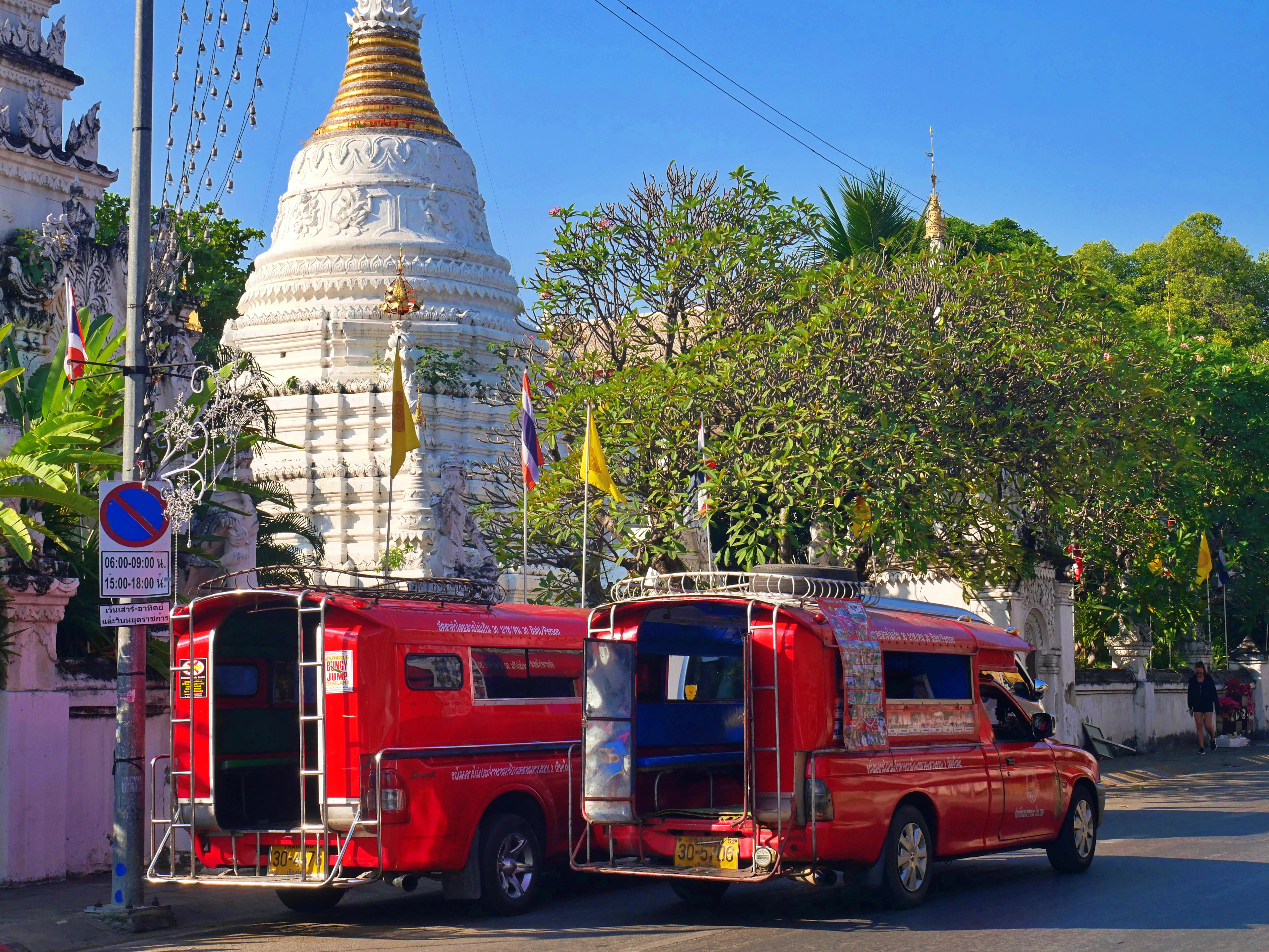 Two red songthaew shared taxis parked at the kerb outside a whitewashed Chiang Mai temple chedi, its gold spire and yellow Buddhist flags bright against a cloudless blue sky