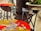 Colorful tables with artistic designs are placed outdoors, featuring intricate iron chairs. One table showcases a whimsical, abstract artwork with vibrant puzzle piece patterns. A nearby chair labeled 'Sweet 45' adds a vintage touch. Large stone planters with green plants provide an earthy contrast.