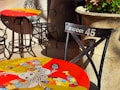 Colorful tables with artistic designs are placed outdoors, featuring intricate iron chairs. One table showcases a whimsical, abstract artwork with vibrant puzzle piece patterns. A nearby chair labeled 'Sweet 45' adds a vintage touch. Large stone planters with green plants provide an earthy contrast.