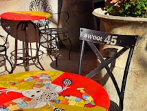 Colorful tables with artistic designs are placed outdoors, featuring intricate iron chairs. One table showcases a whimsical, abstract artwork with vibrant puzzle piece patterns. A nearby chair labeled 'Sweet 45' adds a vintage touch. Large stone planters with green plants provide an earthy contrast.