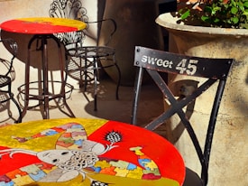 Colorful tables with artistic designs are placed outdoors, featuring intricate iron chairs. One table showcases a whimsical, abstract artwork with vibrant puzzle piece patterns. A nearby chair labeled 'Sweet 45' adds a vintage touch. Large stone planters with green plants provide an earthy contrast.