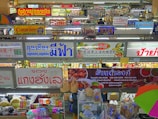 A bustling indoor market scene with multiple shop signs displayed prominently, written in various languages including Thai and Chinese. There are stalls selling a variety of goods, including food and groceries. The image is colorful with items neatly arranged on shelves and hanging from hooks.