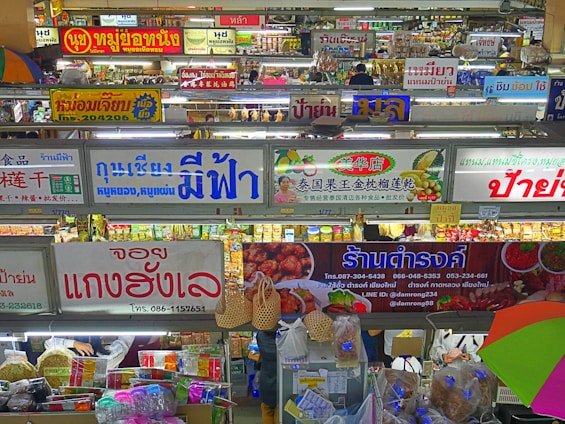 A bustling indoor market scene with multiple shop signs displayed prominently, written in various languages including Thai and Chinese. There are stalls selling a variety of goods, including food and groceries. The image is colorful with items neatly arranged on shelves and hanging from hooks.