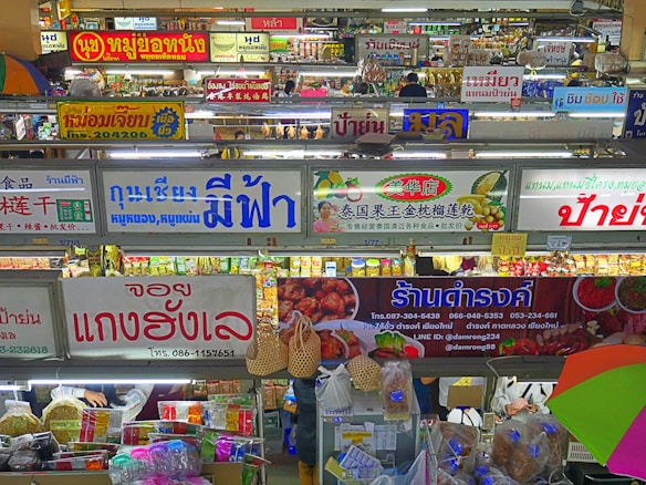 A bustling indoor market scene with multiple shop signs displayed prominently, written in various languages including Thai and Chinese. There are stalls selling a variety of goods, including food and groceries. The image is colorful with items neatly arranged on shelves and hanging from hooks.