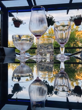 A variety of crystal glasses are neatly arranged on a reflective glass table creating an elegant mirror image. The backdrop consists of lush greenery and hanging potted plants, set against a partly cloudy blue sky.