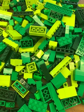 A pile of LEGO bricks in various shades of green and yellow, arranged haphazardly. The pieces vary in size and include standard rectangular bricks and smaller, more specialized shapes.