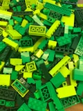 A pile of LEGO bricks in various shades of green and yellow, arranged haphazardly. The pieces vary in size and include standard rectangular bricks and smaller, more specialized shapes.