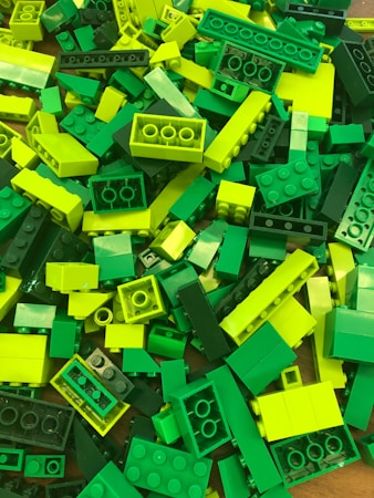 A pile of LEGO bricks in various shades of green and yellow, arranged haphazardly. The pieces vary in size and include standard rectangular bricks and smaller, more specialized shapes.