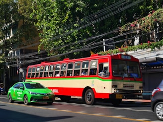 A colorful, vintage bus is moving along a street, accompanied by a modern green taxi. The bus is red and cream-colored with a 65 route sign, while the taxi is vividly green. The scene includes lush green trees and foliage along the sidewalk, with power lines overhead and a building partially visible behind the trees.