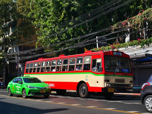 A colorful, vintage bus is moving along a street, accompanied by a modern green taxi. The bus is red and cream-colored with a 65 route sign, while the taxi is vividly green. The scene includes lush green trees and foliage along the sidewalk, with power lines overhead and a building partially visible behind the trees.