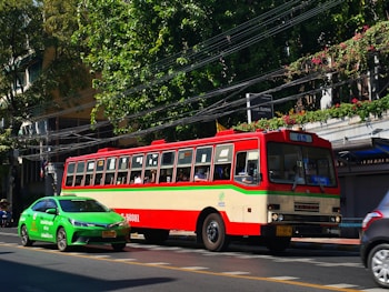 A colorful, vintage bus is moving along a street, accompanied by a modern green taxi. The bus is red and cream-colored with a 65 route sign, while the taxi is vividly green. The scene includes lush green trees and foliage along the sidewalk, with power lines overhead and a building partially visible behind the trees.