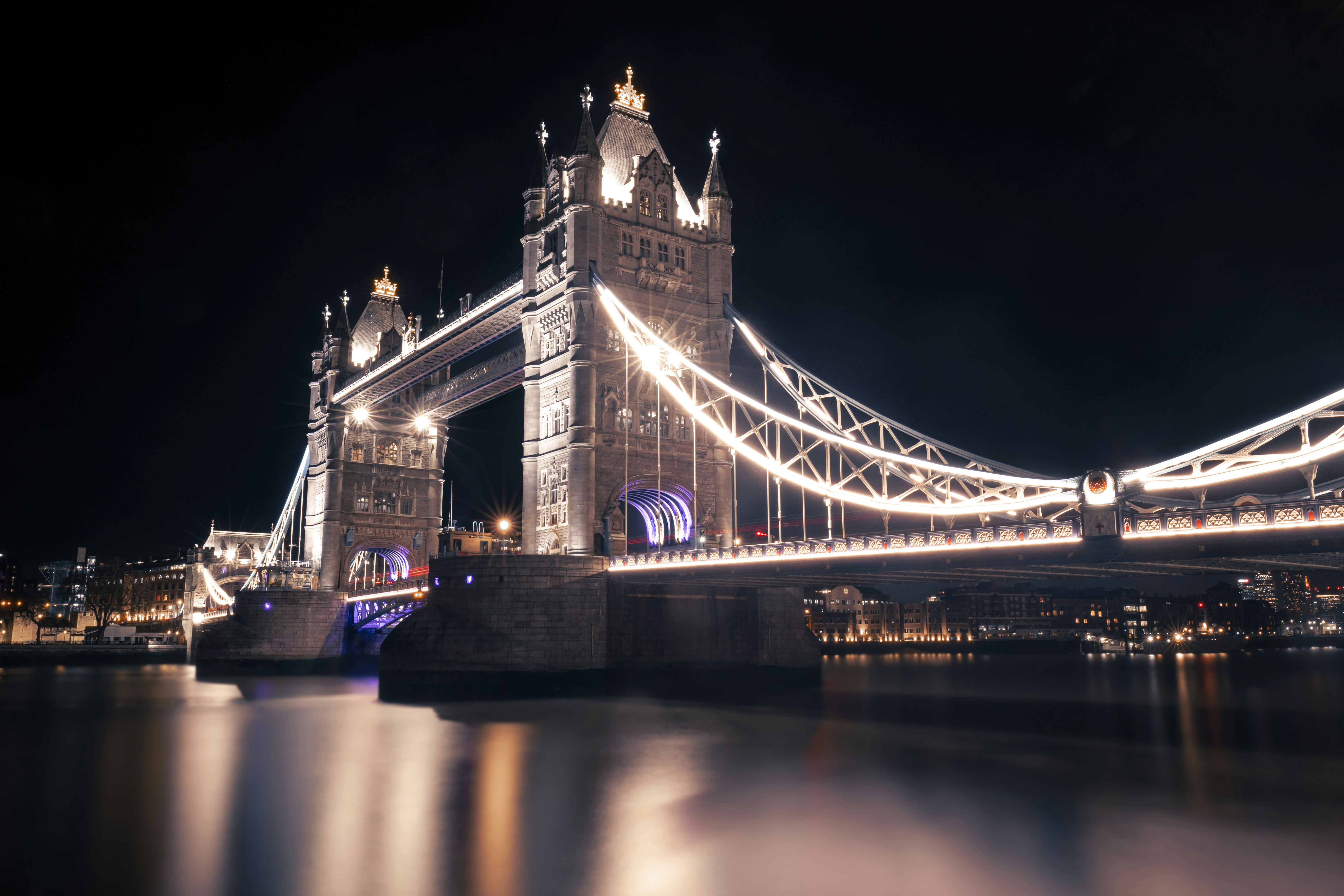 London cityscape at night with illuminated Tower Bridge and city lights reflecting in the Thames, deep dark blue sky, dramatic atmospheric lighting