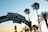 A vintage-style sign arching over an entrance reads 'Santa Monica Yacht Harbor Sport Fishing Boating Cafes' with palm trees silhouetted against a clear blue sky. The sun is setting, casting warm golden hues.