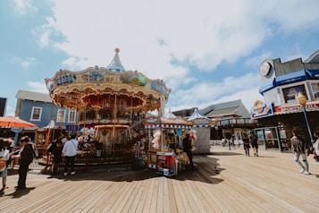 A vibrant carousel decorated with colorful lights and intricate designs stands on a wooden boardwalk. Several food stalls are nearby, offering snacks like churros. People are walking around enjoying the fairground atmosphere. Blue skies with scattered clouds add to the lively setting.