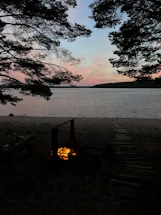 A serene lakeside campsite at dusk with a glowing campfire and dense forest in the background.