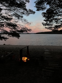 A cozy RV parked beside a serene lake at sunset, with camping chairs and a small campfire glowing warmly.