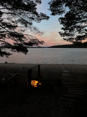 A serene lakeside campsite at sunset with a glowing campfire and silhouetted pine trees.