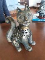 A decorative cat sculpture made of metal with intricate cutout designs on its body, featuring a swirling tail and artistic facial details. It is positioned on a wooden surface with a blurred background of furniture, including a blue upholstered chair.