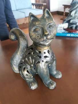 A decorative cat sculpture made of metal with intricate cutout designs on its body, featuring a swirling tail and artistic facial details. It is positioned on a wooden surface with a blurred background of furniture, including a blue upholstered chair.