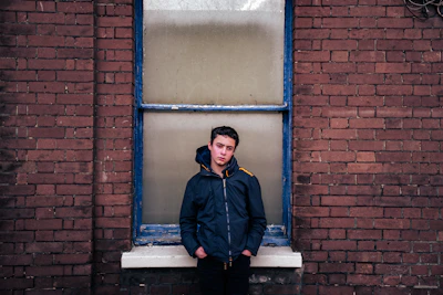 A young person wearing a sleek, understated jacket standing against a soft pastel wall.