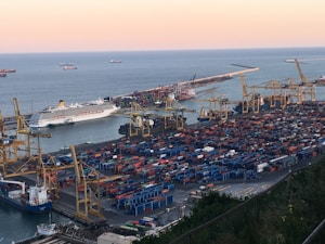 A bustling port with numerous shipping containers organized in neat rows, several large cranes positioned around, and a few ships docked by the pier. The sea stretches into the distance with additional ships visible on the horizon under a soft, pastel sky.