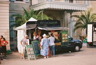 A food truck is set up beside a historical building, with palm trees and a beige stone facade. Several people are gathered around the truck, reading menus and placing orders. The setup includes a canopy tent for shade and menu boards displaying colorful food images.