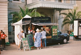 A food truck is set up beside a historical building, with palm trees and a beige stone facade. Several people are gathered around the truck, reading menus and placing orders. The setup includes a canopy tent for shade and menu boards displaying colorful food images.
