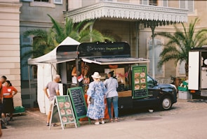 A food truck is set up beside a historical building, with palm trees and a beige stone facade. Several people are gathered around the truck, reading menus and placing orders. The setup includes a canopy tent for shade and menu boards displaying colorful food images.