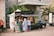 A food truck is set up beside a historical building, with palm trees and a beige stone facade. Several people are gathered around the truck, reading menus and placing orders. The setup includes a canopy tent for shade and menu boards displaying colorful food images.
