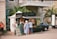 A food truck is set up beside a historical building, with palm trees and a beige stone facade. Several people are gathered around the truck, reading menus and placing orders. The setup includes a canopy tent for shade and menu boards displaying colorful food images.