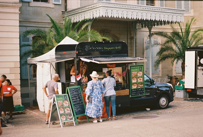 A food truck is set up beside a historical building, with palm trees and a beige stone facade. Several people are gathered around the truck, reading menus and placing orders. The setup includes a canopy tent for shade and menu boards displaying colorful food images.