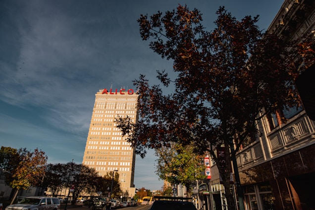 A tall building with the word 'ALICO' in red letters stands prominently against a clear sky. The building is surrounded by smaller structures and trees with autumn foliage. The scene is urban with vehicles parked along the street, creating a sense of a bustling city environment.
