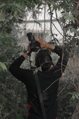 A person is standing in a dense thicket of green foliage, holding a camera with a large lens aimed upwards. The camera strap hangs over their shoulder, and they are wearing a dark jacket with red accents. The scene is surrounded by intertwined branches and leaves.