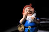A colorful garden gnome with a cheerful expression stands against a dark background. The gnome is wearing a bright blue outfit and a red hat, with a white beard. It is holding its cheek with one hand and has a small wooden birdhouse hanging on its belt.