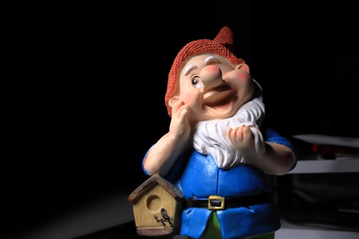 A colorful garden gnome with a cheerful expression stands against a dark background. The gnome is wearing a bright blue outfit and a red hat, with a white beard. It is holding its cheek with one hand and has a small wooden birdhouse hanging on its belt.
