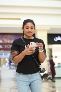 A person wearing a black shirt with patterned sleeves and blue jeans is looking at a smartphone. They are carrying a small black crossbody bag and standing in an indoor shopping area with blurred shops in the background.