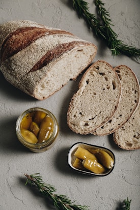 A rustic loaf of bread with a crusty texture is partially sliced on a textured surface. Nearby, a jar and a small dish contain pickled peppers. Fresh rosemary sprigs are placed around the setup, adding a touch of greenery.
