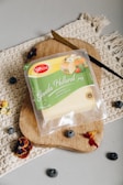 A package of Gouda Holland cheese rests on a wooden cutting board. The packaging is green and transparent, showing slices of cheese with a label. Surrounding the board are a few scattered blueberries and dried flower petals, all placed atop a textured fabric.