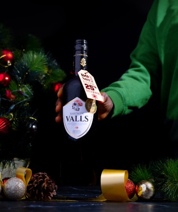 A hand dressed in a green sleeve holds a bottle of wine labeled 'Baron de Valls.' The bottle has a tag indicating a discount until December 25th. The setting includes holiday decorations such as pine branches, pine cones, and red and gold ornaments. The background is dark, highlighting the festive elements.