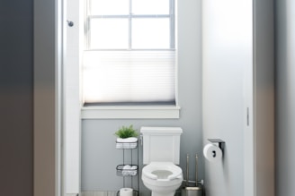 A small bathroom with a toilet situated under a window partially covered by a blind. A stand next to the toilet holds toilet paper and a small potted plant. The walls are painted light grey, and the room is well-lit by natural light coming through the window.