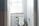 A small bathroom with a toilet situated under a window partially covered by a blind. A stand next to the toilet holds toilet paper and a small potted plant. The walls are painted light grey, and the room is well-lit by natural light coming through the window.