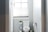 A small bathroom with a toilet situated under a window partially covered by a blind. A stand next to the toilet holds toilet paper and a small potted plant. The walls are painted light grey, and the room is well-lit by natural light coming through the window.
