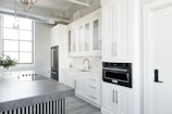 A modern kitchen features sleek white cabinetry, a built-in oven, and a large countertop island with a stovetop. The space is brightly lit by a large window, and a vase with green foliage sits on the counter. Stainless steel appliances add to the contemporary feel.