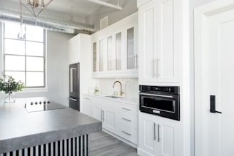 A modern kitchen features sleek white cabinetry, a built-in oven, and a large countertop island with a stovetop. The space is brightly lit by a large window, and a vase with green foliage sits on the counter. Stainless steel appliances add to the contemporary feel.
